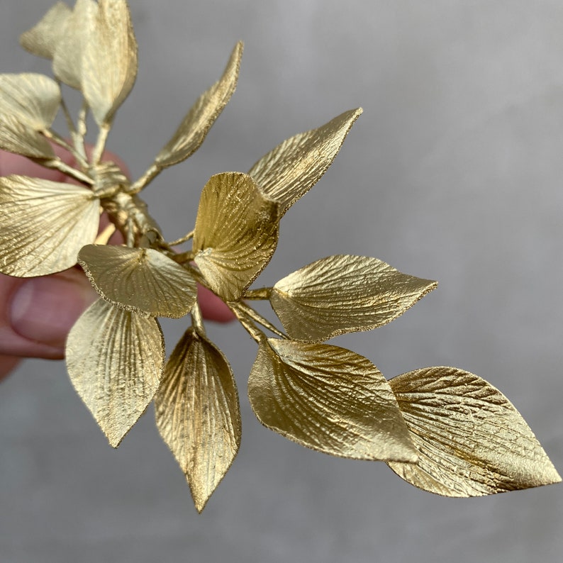May include: A gold-colored hair accessory featuring a branch-like design with multiple detailed leaves. The leaves have a textured surface and are arranged in a natural, organic pattern. The accessory is likely for bridal or formal wear.