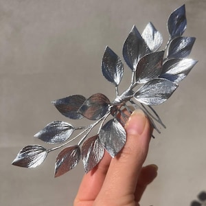 May include: Silver-colored hair comb featuring a branch design with multiple detailed leaves. The leaves have a textured appearance, and the comb is held against a neutral background. A decorative hair accessory.