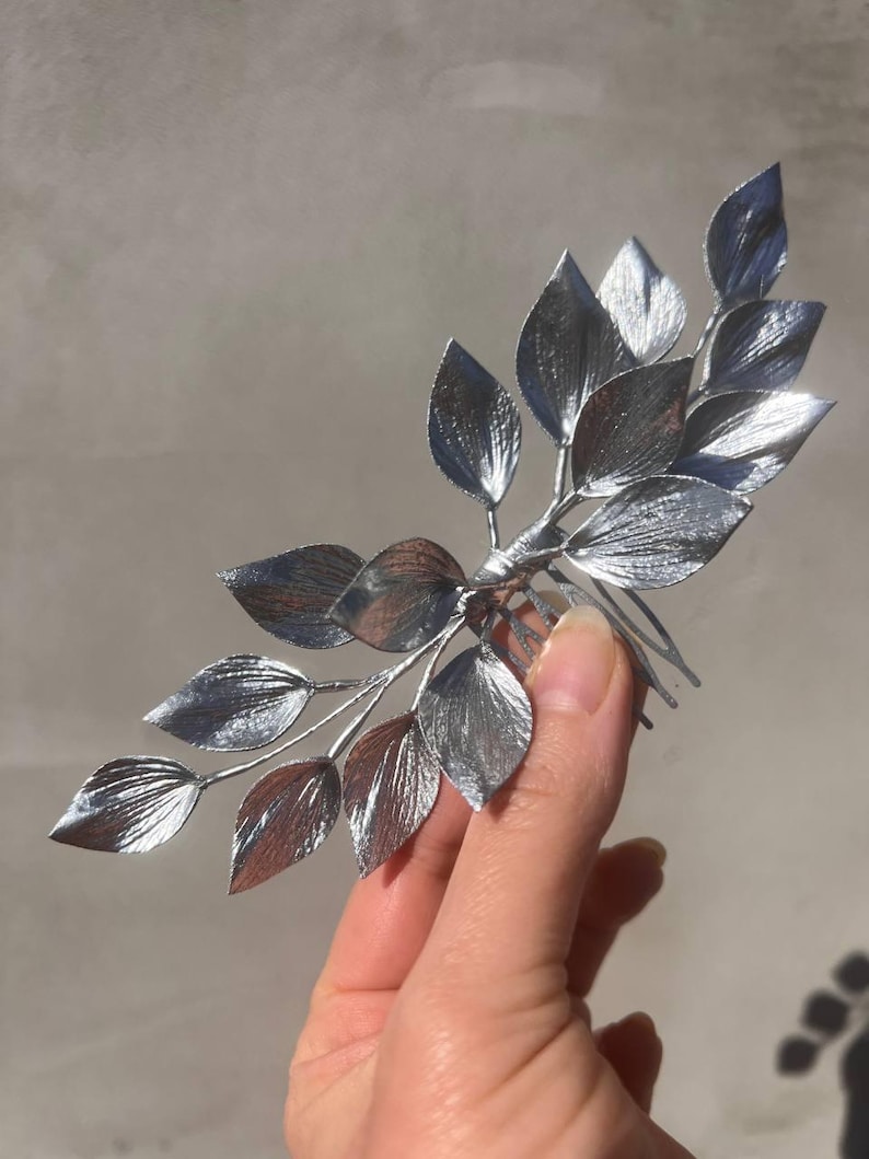 May include: Silver-colored hair comb featuring a branch design with multiple detailed leaves. The leaves have a textured appearance, and the comb is held against a neutral background. A decorative hair accessory.