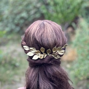May include: A gold-colored leaf hair comb is set in dark brown hair. The hair accessory is made of metal and features several detailed leaves. The background is a blurred view of green foliage.