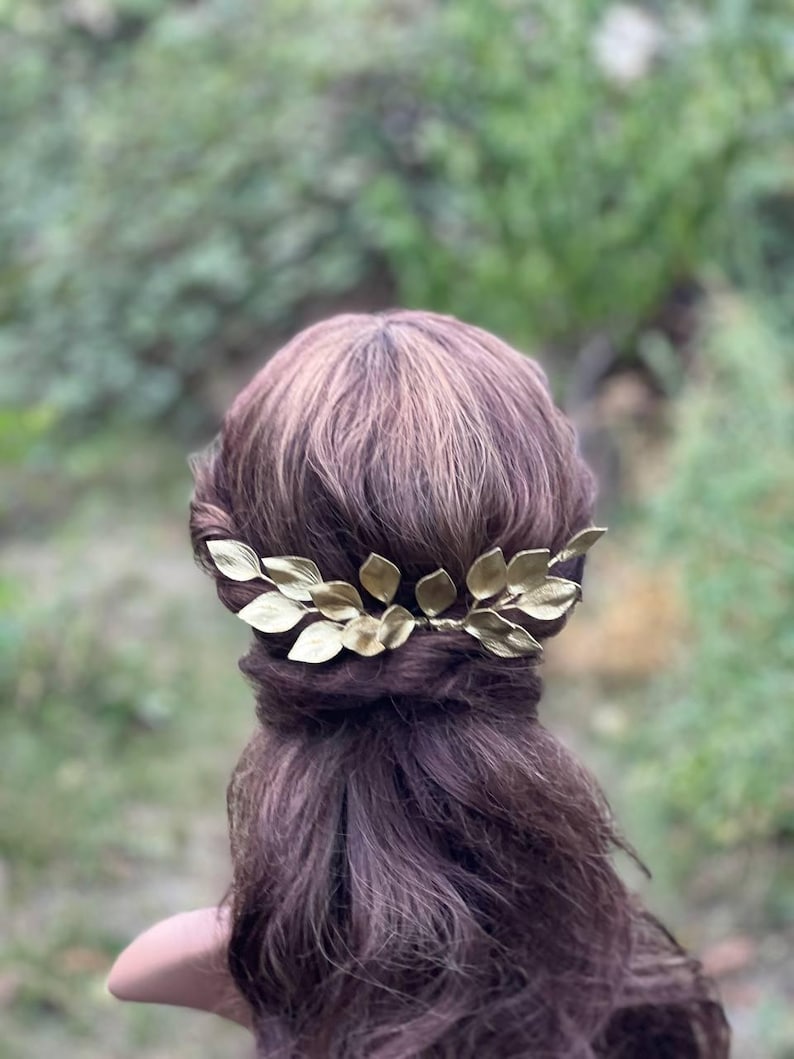 May include: A gold-colored leaf hair comb is set in dark brown hair. The hair accessory is made of metal and features several detailed leaves. The background is a blurred view of green foliage.