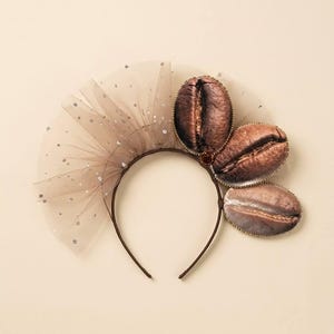 May include: A headband featuring three oval-shaped coffee bean decorations on one side and a beige tulle fan with silver glitter accents on the other. The coffee bean embellishments are brown with realistic textures and gold-toned beaded edges.