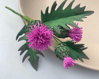 Puede incluir: Un ramo de tres flores de cardo púrpura con hojas verdes. Las flores están hechas de papel y tienen un aspecto realista.