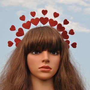 May include: A red glitter heart headband with a row of hearts around the crown. The headband is worn by a mannequin with long brown hair.