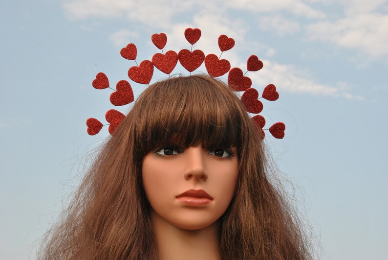 May include: A red glitter heart headband with a row of hearts around the crown. The headband is worn by a mannequin with long brown hair.