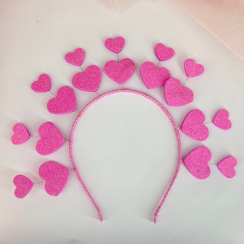May include: Pink glitter heart headband with a silver headband. The headband is decorated with multiple pink glitter hearts.