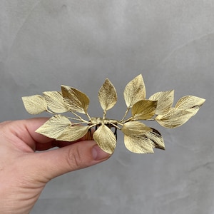 May include: A gold-colored hair comb featuring a leaf design. The comb has multiple leaves arranged in a symmetrical pattern. The leaves have a textured surface and are attached to a comb base.