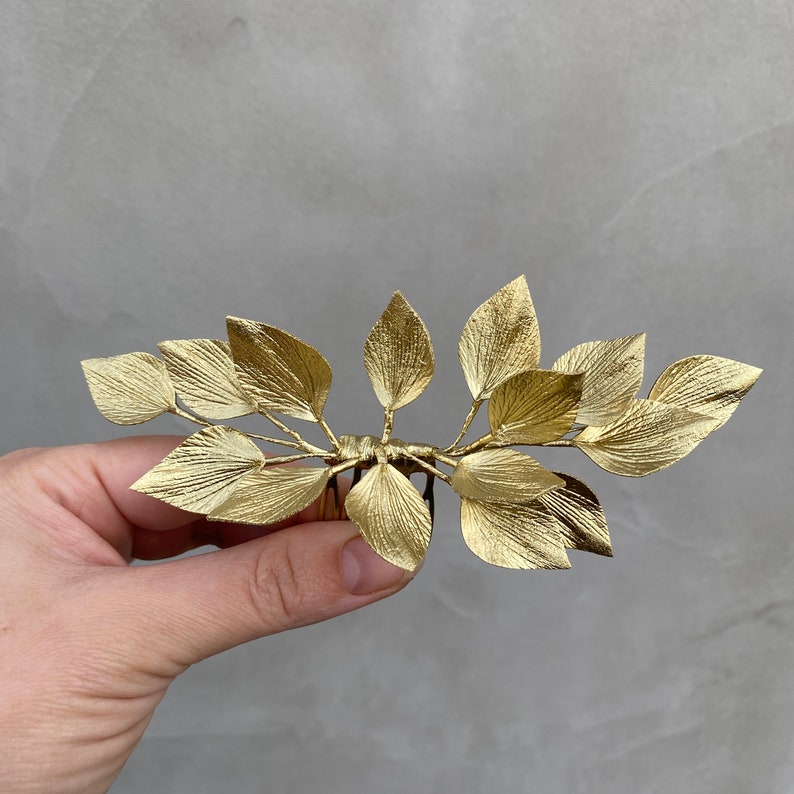 May include: A gold-colored hair comb featuring a leaf design. The comb has multiple leaves arranged in a symmetrical pattern. The leaves have a textured surface and are attached to a comb base.