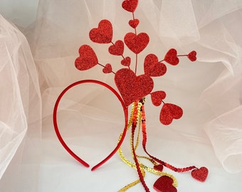 May include: Red heart-shaped headband with a gold sequined tail. The headband is decorated with a cluster of red hearts.