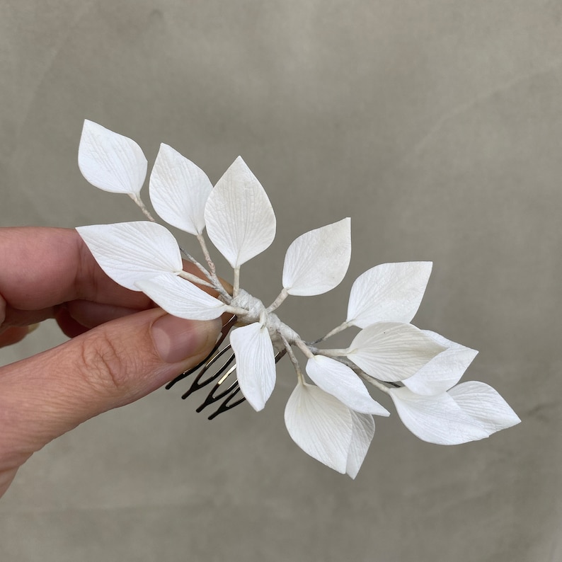 May include: A white leaf hair comb. The comb features delicate, textured white leaves arranged on a flexible wire base. The leaves are attached to a black comb. The design is suitable for weddings or special occasions.