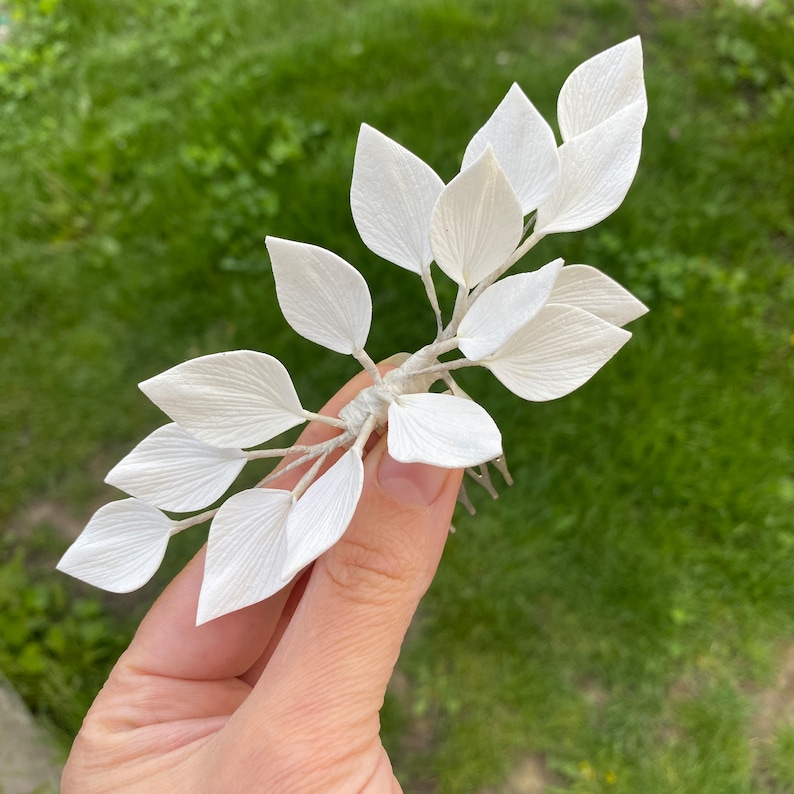 May include: A delicate white floral hair comb featuring multiple textured leaves. The leaves are arranged on a slender, flexible stem, and attached to a metal comb. The comb is designed for securing the hair and adding a touch of elegance.