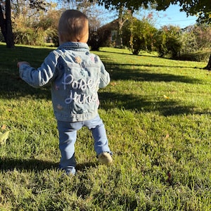 May include: A toddler wearing a light blue denim jacket with colorful embroidered flowers and the text "Posy Jane" on the back. The child is standing on a grassy lawn.