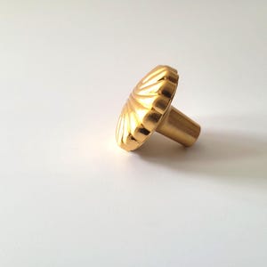 May include: A gold-coloured cabinet knob with a decorative, fan-shaped design. The knob has a cylindrical stem for mounting and is set against a white background. Suitable for drawers and cabinets.