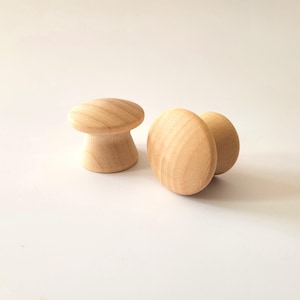 May include: Three unfinished wooden cabinet knobs. One is mushroom-shaped, the other two are oval. The knobs are made of light-coloured wood and are set against a white background.