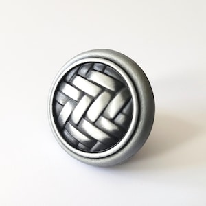 May include: A circular cabinet knob in a silver tone, featuring a woven basket pattern at its centre. The knob has a raised, circular edge and is set against a white backdrop. The knob is likely made of metal.