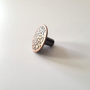 Peut inclure: Un bouton de meuble décoratif avec une face circulaire texturée de couleur cuivre et une tige noire. La face présente un motif organique détaillé. Le bouton est conçu pour les meubles et la décoration intérieure.