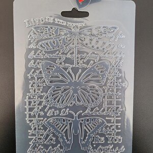 May include: A clear plastic craft mould featuring dragonfly and butterfly designs, with script reading "Live in the Moment" and "Be You". The mould has a grid pattern and additional script elements.