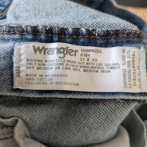 Puede incluir: Primer plano de una etiqueta de vaqueros Wrangler. La etiqueta blanca con texto negro muestra la marca, la talla 31 x 30 y las instrucciones de cuidado. Los vaqueros son de color denim azul claro.