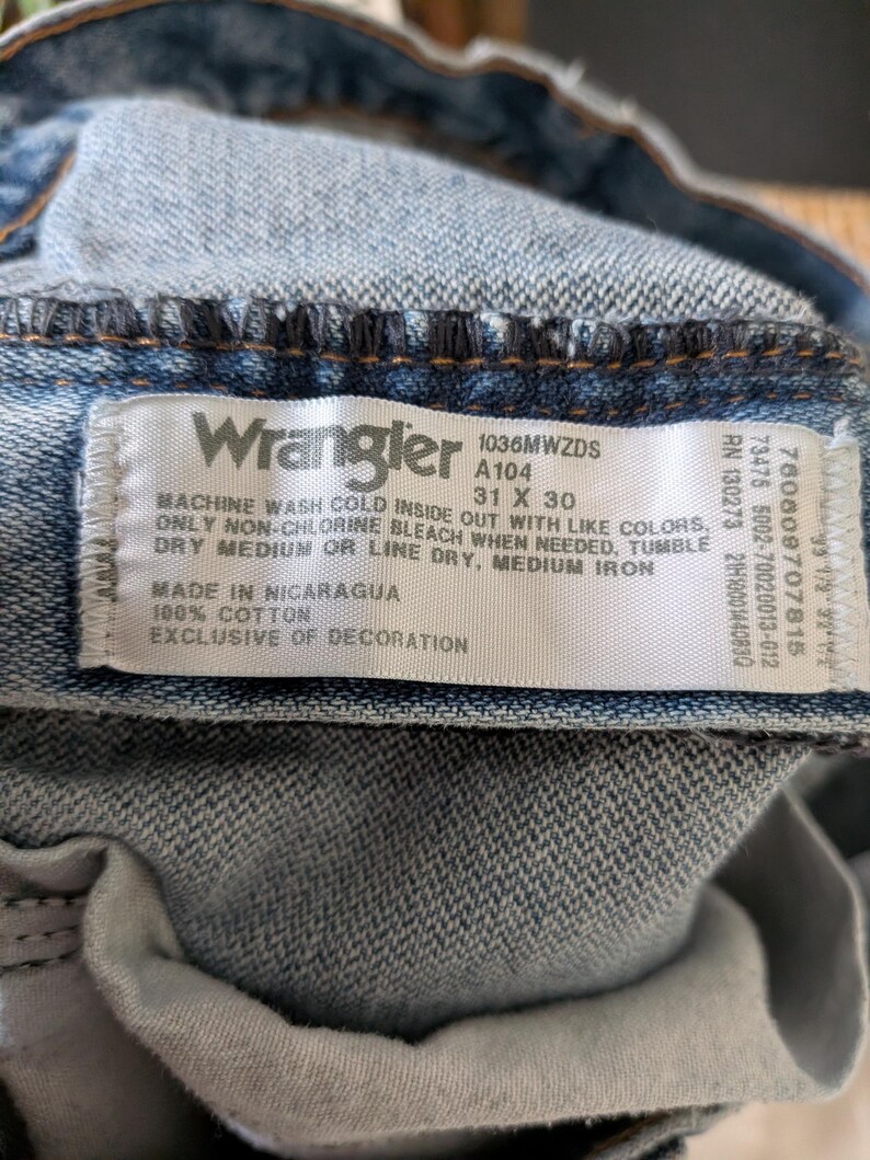 Puede incluir: Primer plano de una etiqueta de vaqueros Wrangler. La etiqueta blanca con texto negro muestra la marca, la talla 31 x 30 y las instrucciones de cuidado. Los vaqueros son de color denim azul claro.
