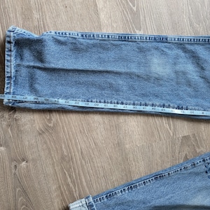 Puede incluir: Vaqueros de mezclilla azul claro con una cinta m&eacute;trica que muestra la medida de la cintura. Los vaqueros est&aacute;n colocados sobre una superficie de madera, mostrando la textura y el color de la tela. Los vaqueros tienen un parche en la parte inferior.