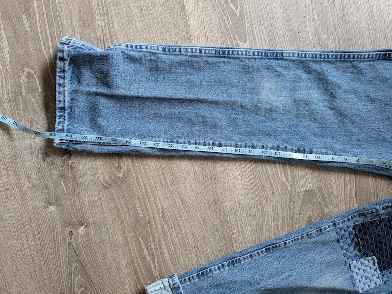 Puede incluir: Vaqueros de mezclilla azul claro con una cinta m&eacute;trica que muestra la medida de la cintura. Los vaqueros est&aacute;n colocados sobre una superficie de madera, mostrando la textura y el color de la tela. Los vaqueros tienen un parche en la parte inferior.