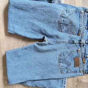 Puede incluir: Vaqueros de mezclilla azul claro con costuras Wrangler cl&aacute;sicas en los bolsillos traseros. Los vaqueros tienen un parche de cuero marr&oacute;n y remaches de lat&oacute;n. Los vaqueros est&aacute;n doblados sobre una superficie de madera clara.