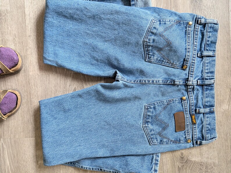 Puede incluir: Vaqueros de mezclilla azul claro con costuras Wrangler cl&aacute;sicas en los bolsillos traseros. Los vaqueros tienen un parche de cuero marr&oacute;n y remaches de lat&oacute;n. Los vaqueros est&aacute;n doblados sobre una superficie de madera clara.