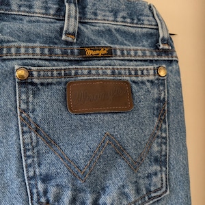 Puede incluir: Primer plano de unos vaqueros azules con una etiqueta Wrangler en el bolsillo trasero. Los vaqueros tienen un dise&ntilde;o cl&aacute;sico de cinco bolsillos, con remaches de lat&oacute;n y un parche de cuero marr&oacute;n. Las costuras son de color beige contrastante.
