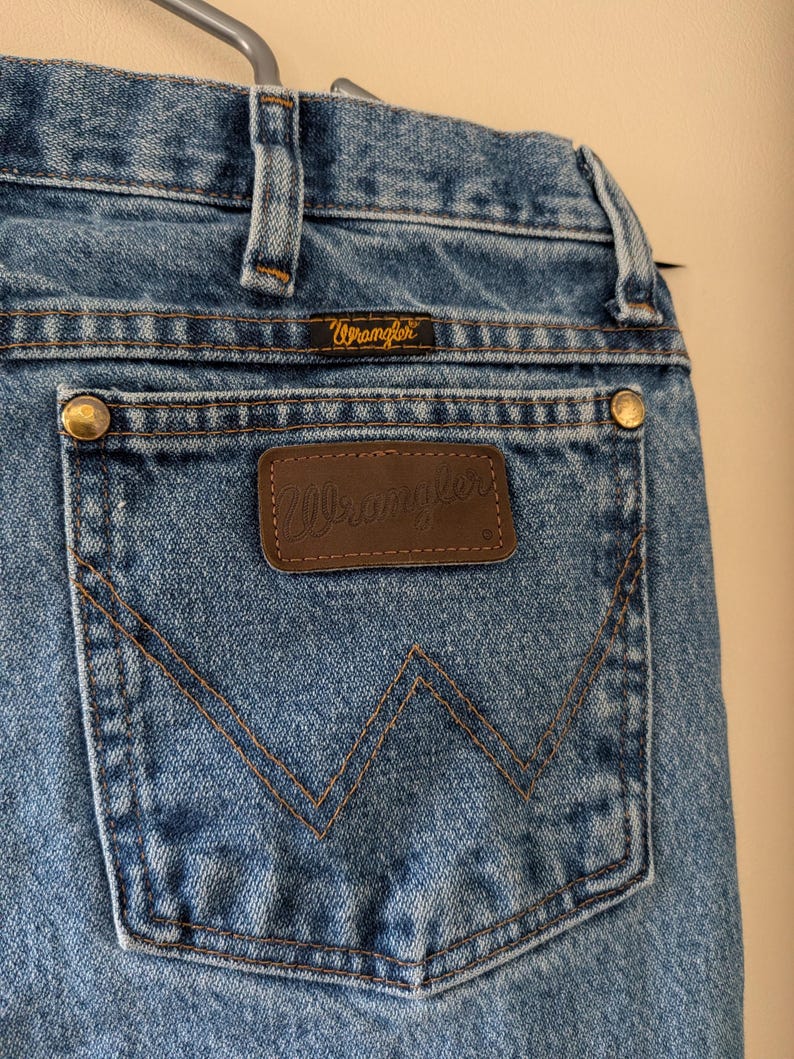 Puede incluir: Primer plano de unos vaqueros azules con una etiqueta Wrangler en el bolsillo trasero. Los vaqueros tienen un dise&ntilde;o cl&aacute;sico de cinco bolsillos, con remaches de lat&oacute;n y un parche de cuero marr&oacute;n. Las costuras son de color beige contrastante.