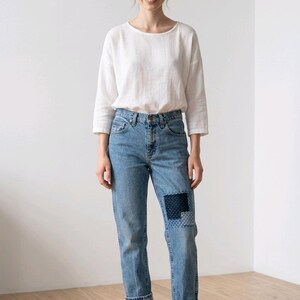 Puede incluir: Una persona viste una camisa blanca de manga larga y vaqueros azul claro con un parche en la rodilla. Tambi&eacute;n lleva mocasines de cuero marr&oacute;n. El atuendo es informal y c&oacute;modo.