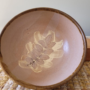 Puede incluir: Cuenco de cerámica hecho a mano, interior marrón claro y borde marrón oscuro. El cuenco presenta un diseño de hojas en tonos beige y marrón. Ideal para servir alimentos o como pieza decorativa. Diámetro: aproximadamente 20 cm.