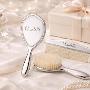 May include: A baby grooming set with a silver-toned brush, comb, and mirror, each engraved with the name "Charlotte." The set includes a soft-bristled brush, a comb, and a hand mirror. A gift box with a ribbon is also present.