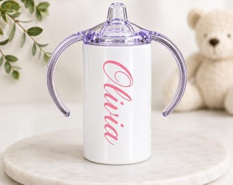 May include: A white sippy cup with two purple handles and a matching purple lid. The name Olivia is printed vertically on the front of the cup in a pink cursive script font. The cup features a smooth cylindrical body and a clear plastic spout.