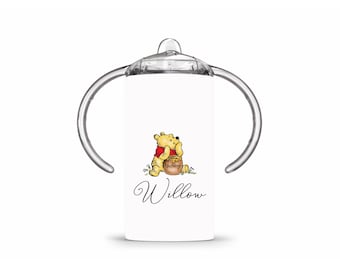 May include: A white baby sippy cup with clear handles and a clear top. The cup features a Winnie the Pooh graphic with a honey pot and the name "Willow" in script.