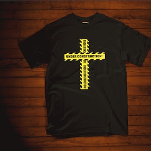 Puede incluir: Camiseta negra con un diseño de cruz amarilla y negra. La cruz está hecha de cinta de precaución con el texto "Under Construction" repetido en cada lado.