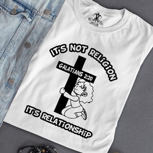 May include: White t-shirt with black text that reads "It's not religion, it's relationship."  The text is in a circle around a black cross with a small cartoon girl hugging the cross. The text "Galatians 2:20" is written on the cross.