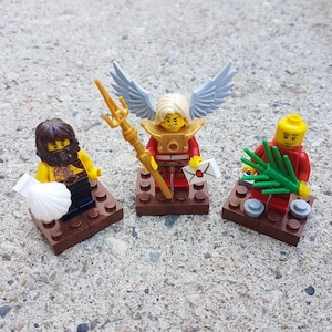 Könnte beinhalten: Drei Lego-Minifiguren, eine mit einer weißen Muschel, eine mit Flügeln und einem goldenen Stab und eine mit einer grünen Pflanze. Die Figuren stehen auf braunen Lego-Grundplatten.