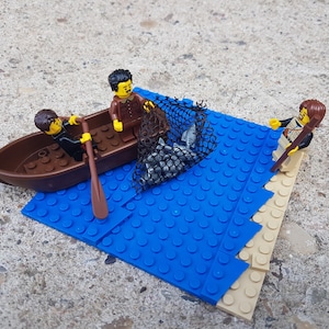 May include: Two Lego figures in a brown boat with oars, one figure is holding a net with grey fish inside. A third Lego figure stands on a blue and tan Lego base, holding a fishing rod.
