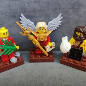 Peut inclure: Trois figurines LEGO debout sur des socles en briques marron. La figurine de gauche porte du rouge et tient une branche de palmier verte. Celle du centre a des ailes blanches, un plastron doré, un trident doré et une enveloppe blanche. La figurine de droite a une barbe brune et tient un coquillage blanc.