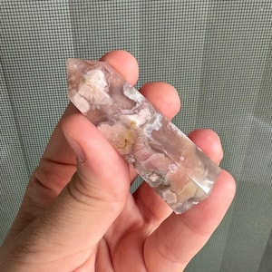 May include: A hand holds a polished crystal point. The crystal features a mix of translucent and opaque sections with a blend of pink, white, and tan colors. The crystal is approximately 4 inches long.