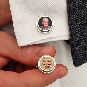 May include: A pair of silver cufflinks. One cufflink features a photo of a man in a round frame. The other cufflink has a wooden surface with the text "Forever by your side" engraved in dark brown.