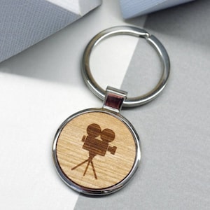 May include: A silver key chain with a round wooden charm featuring a laser-engraved silhouette of a vintage movie camera.