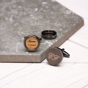 May include: Two cufflinks, one with a wood background and the text "Happy 5 years" engraved in gold, and the other with a black background and the initials "C.F." engraved in gold.