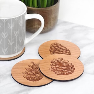 May include: Set of three round wooden coasters with a brown pine cone design. The coasters are made of natural wood and have a light brown color.
