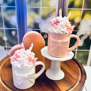May include: Two decorative mugs, one pink and one white, each topped with whipped cream, pink bows, and straws. The mugs have bow designs. The pink mug sits on a white pedestal, while the white mug rests on a wooden tray.