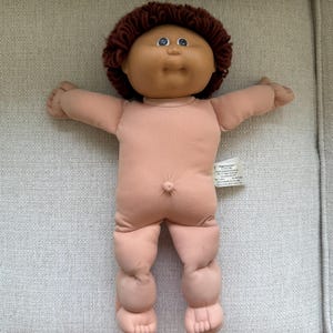 May include: A Cabbage Patch Kids doll with a soft, light-toned fabric body, a vinyl head, and dark brown yarn hair. The doll features a round face, blue eyes, and a small belly button. A white fabric tag is attached to the side of the torso.