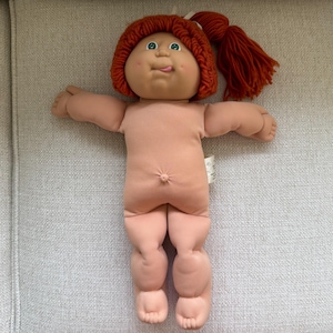 May include: A soft-bodied doll with a light peach-colored fabric skin, a prominent belly button, and green eyes. The doll features a round face with a small tongue sticking out and thick, textured, reddish-brown yarn hair tied in a side ponytail.