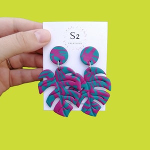 May include: A pair of pink and teal dangle earrings featuring a monstera leaf design. The earrings are made of polymer clay and have silver hooks.