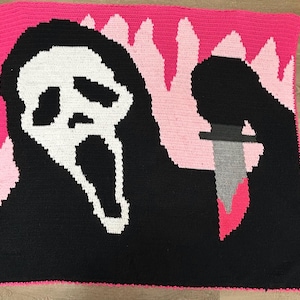 May include: A handmade crochet blanket featuring a black and white Ghostface mask from the Scream horror film series. The blanket has a pink border and a pink and light pink background with a knife. The blanket is a unique, handmade item.