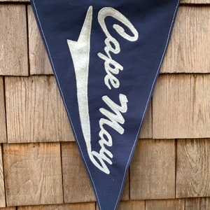May include: A blue triangular pennant with white embroidered text that reads "Cape May" in a cursive font. The pennant is hanging on a wooden wall.